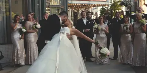 Cinematic wedding scene of a bride and groom kissing on a city street while the wedding party cheers and holds bouquets.