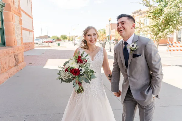 A bride holding a bouquet of red and white flowers and a groom in a grey suit walking hand-in-hand on a city sidewalk.