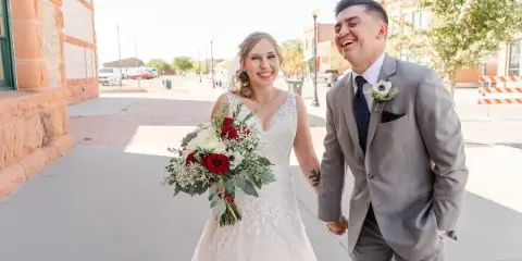A bride holding a bouquet of red and white flowers and a groom in a grey suit walking hand-in-hand on a city sidewalk.
