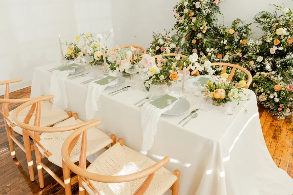 A wedding reception table set with white linens, green place settings, and floral centerpieces, surrounded by light wood accent chairs with woven seats.