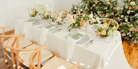 A wedding reception table set with white linens, green place settings, and floral centerpieces, surrounded by light wood accent chairs with woven seats.