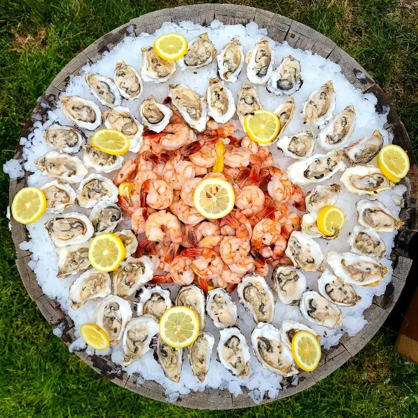 A large circular wooden platter filled with ice, topped with a ring of raw oysters and a central mound of cooked shrimp, garnished with lemon wedges.