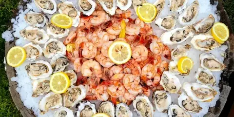 A large circular wooden platter filled with ice, topped with a ring of raw oysters and a central mound of cooked shrimp, garnished with lemon wedges.