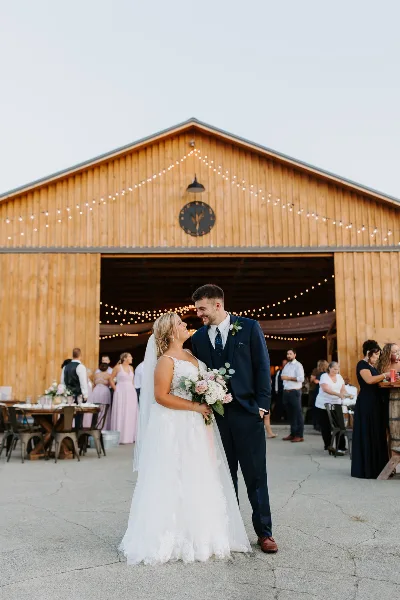 A bride and groom standing in front of a rustic wooden barn wedding venue with string lights and guests in the background.