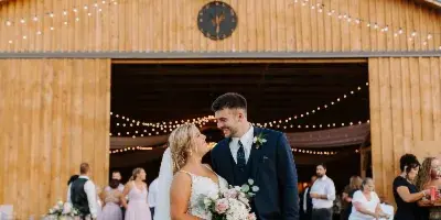 A bride and groom standing in front of a rustic wooden barn wedding venue with string lights and guests in the background.