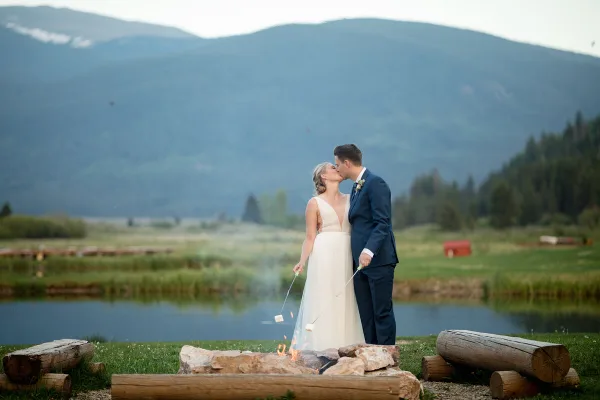 A bride and groom kissing outdoors near a stone fire pit with a mountain and lake background.