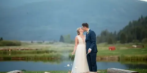 A bride and groom kissing outdoors near a stone fire pit with a mountain and lake background.