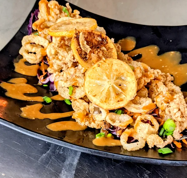 A close-up of a plated dish with fried seafood, lemon slices, and sauce on a black plate.