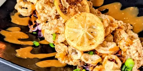 A close-up of a plated dish with fried seafood, lemon slices, and sauce on a black plate.