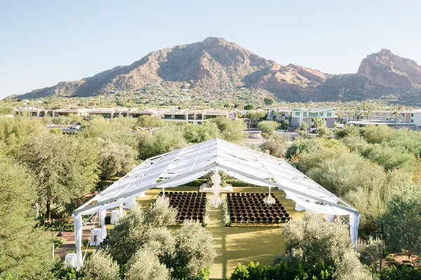 Aerial view of a white event tent with rows of chairs and chandeliers set up in a desert landscape with mountains in the background.