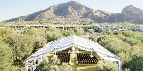 Aerial view of a white event tent with rows of chairs and chandeliers set up in a desert landscape with mountains in the background.