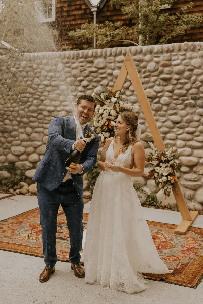 A bride and groom celebrating with a champagne bottle in front of a floral wedding arch.