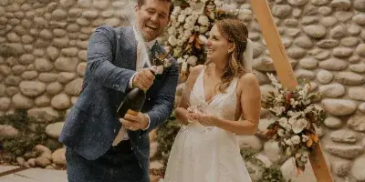 A bride and groom celebrating with a champagne bottle in front of a floral wedding arch.