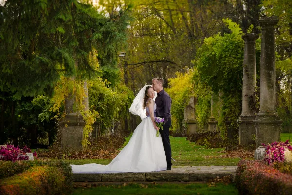 A bride and groom embracing in a garden setting with stone pillars and autumn foliage.