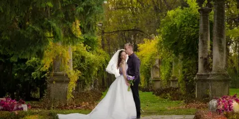 A bride and groom embracing in a garden setting with stone pillars and autumn foliage.