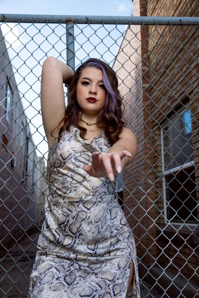 A professional portrait of a young woman with purple hair wearing a snakeskin dress, posing in front of a chain-link fence and brick building.