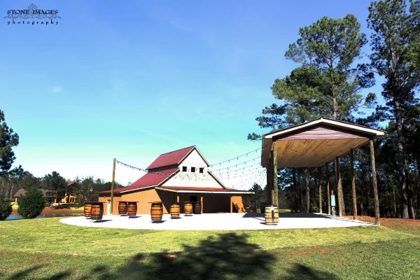 A wide shot of a farm venue featuring a barn-style building with a red roof and an adjacent open-air pavilion with string lights, set on a grassy lawn.