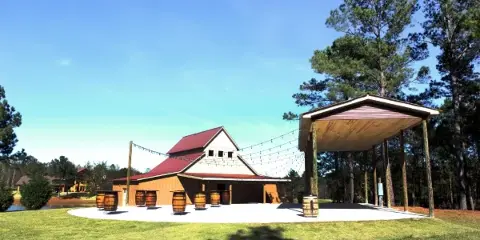 A wide shot of a farm venue featuring a barn-style building with a red roof and an adjacent open-air pavilion with string lights, set on a grassy lawn.