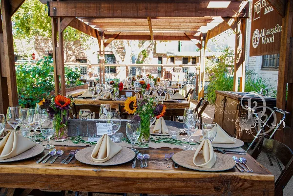 A long wooden table set for an event with chairs, napkins, glassware, and floral centerpieces under a wooden pergola.