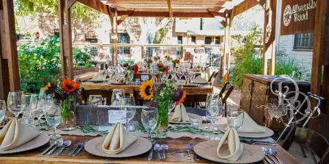A long wooden table set for an event with chairs, napkins, glassware, and floral centerpieces under a wooden pergola.