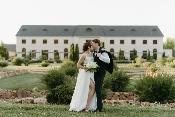 Bride and groom kissing on a lawn in front of a white wedding venue building.