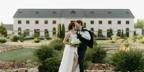 Bride and groom kissing on a lawn in front of a white wedding venue building.