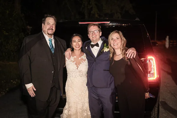 A bride, groom, and two guests posing in front of a black SUV at night.