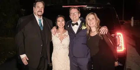 A bride, groom, and two guests posing in front of a black SUV at night.