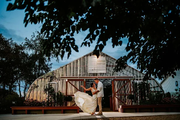 A bride and groom dancing in front of a large greenhouse structure at a farm venue.