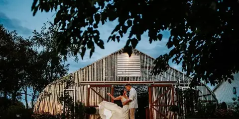 A bride and groom dancing in front of a large greenhouse structure at a farm venue.