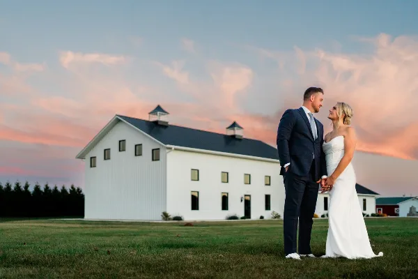 Bride and groom standing on a lawn in front of a white barn-style wedding venue at sunset