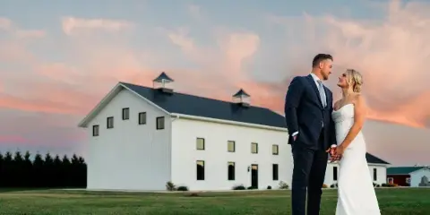 Bride and groom standing on a lawn in front of a white barn-style wedding venue at sunset