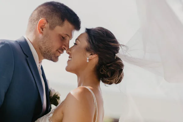 A close-up professional wedding photograph of a bride and groom smiling at each other, with the bride's veil visible in the background.