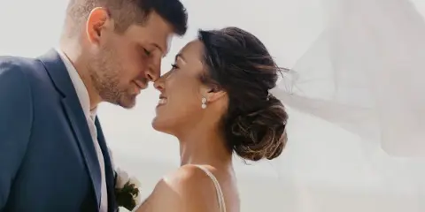 A close-up professional wedding photograph of a bride and groom smiling at each other, with the bride's veil visible in the background.