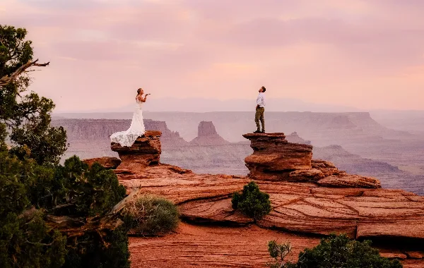 A bride in a white gown and a groom in a white shirt standing on separate red rock formations overlooking a vast canyon at sunset.