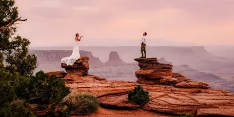 A bride in a white gown and a groom in a white shirt standing on separate red rock formations overlooking a vast canyon at sunset.