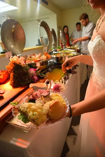 A bride in a white wedding dress holding a plate of food, including steak and bread, while serving herself from a buffet line with chafing dishes and floral decorations.