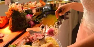 A bride in a white wedding dress holding a plate of food, including steak and bread, while serving herself from a buffet line with chafing dishes and floral decorations.