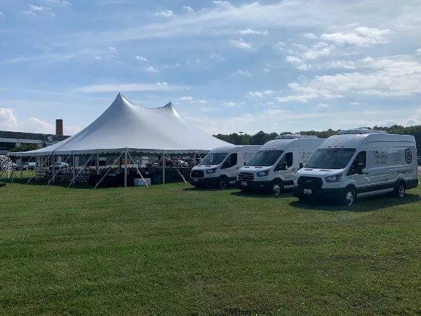 White catering vans with 'Full Service On-Site Catering' branding parked next to a large white event tent on a grassy field.
