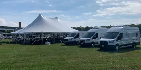White catering vans with 'Full Service On-Site Catering' branding parked next to a large white event tent on a grassy field.