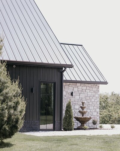 Exterior view of a modern wedding venue building with a dark metal roof, stone wall, and a tiered fountain in the foreground.