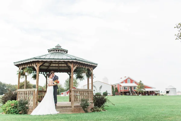 A bride and groom standing in a wooden gazebo on a grassy lawn with a red barn and silo in the background.