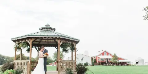 A bride and groom standing in a wooden gazebo on a grassy lawn with a red barn and silo in the background.