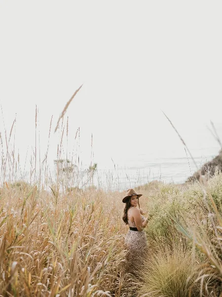 A woman in a hat and dress standing in tall coastal grasses near the ocean.