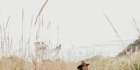 A woman in a hat and dress standing in tall coastal grasses near the ocean.