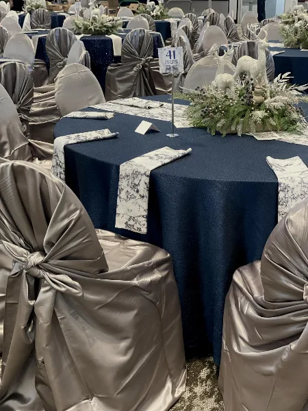 Reception tables set with navy tablecloths, patterned runners, and champagne-colored chair covers with self-tie sashes.
