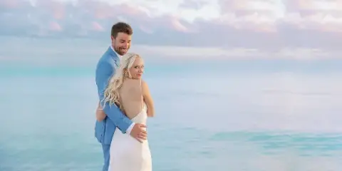 A bride and groom embracing on a beach with a turquoise ocean and pastel sky in the background.