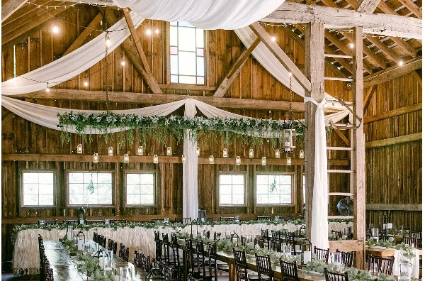 Interior of a rustic wooden barn wedding venue featuring exposed beams, string lights, and a long banquet table set with greenery and candles.