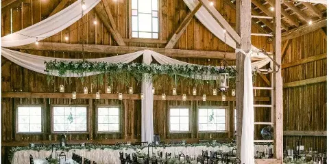 Interior of a rustic wooden barn wedding venue featuring exposed beams, string lights, and a long banquet table set with greenery and candles.