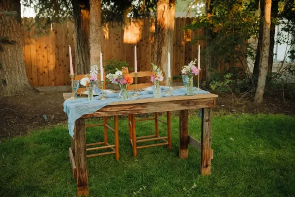 A rustic wooden table set outdoors with a blue linen runner, floral arrangements, and tall candles.